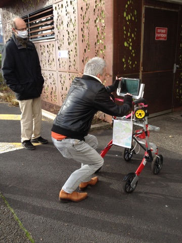 un homme, accroupi devant un chariot, manipule une tablette fixée au sommet de l'engin roulant. À sa gauche, un homme l'observe.